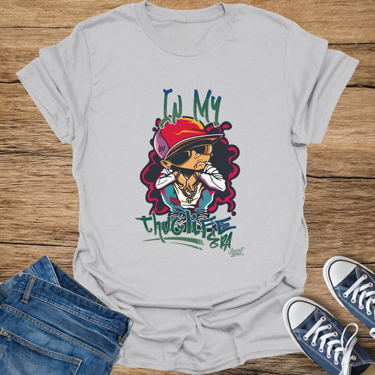 White t-shirt with cartoon of a child in a red hat, sunglasses, jewelry, and text "IN MY THUG LIFE".