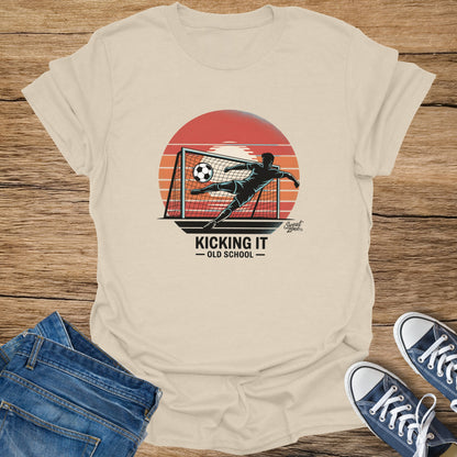 T-shirt with graphic of a soccer player kicking a ball into a net at sunset, text "Kicking It Old School".