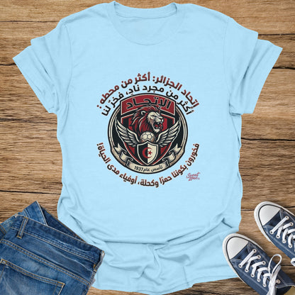 Light blue t-shirt with circular logo featuring a lion, soccer ball, wings, and Arabic text.