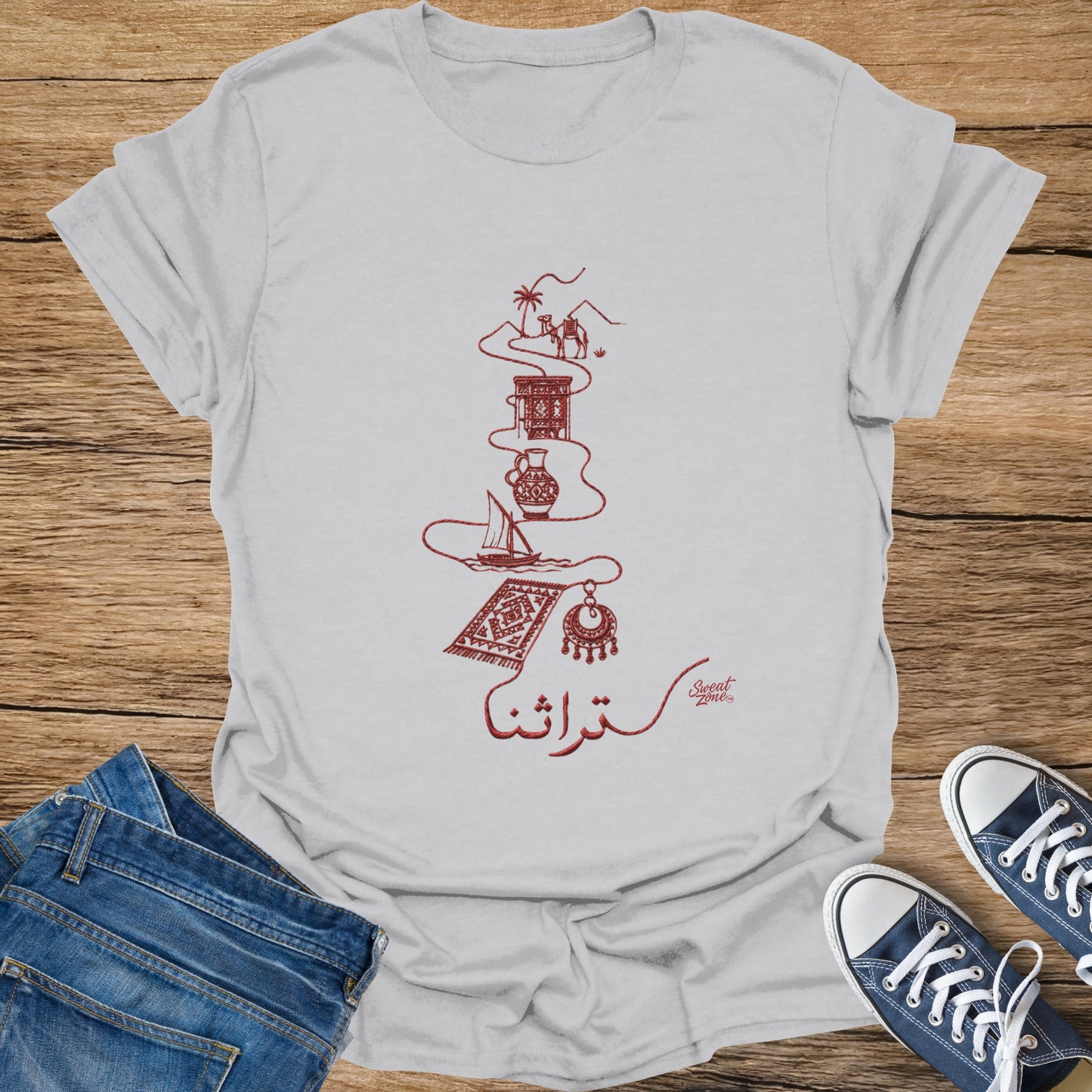 White t-shirt with red embroidery of a camel, pottery, sailboat, rug, and jewelry with Arabic text.