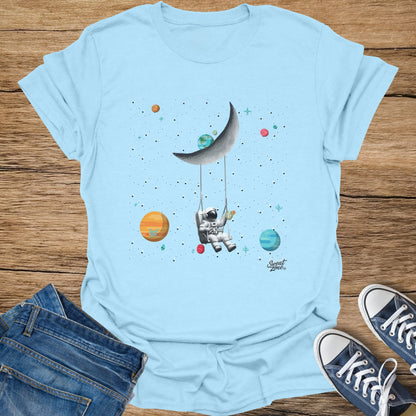 Light blue t-shirt with astronaut swinging on moon, surrounded by planets and stars.