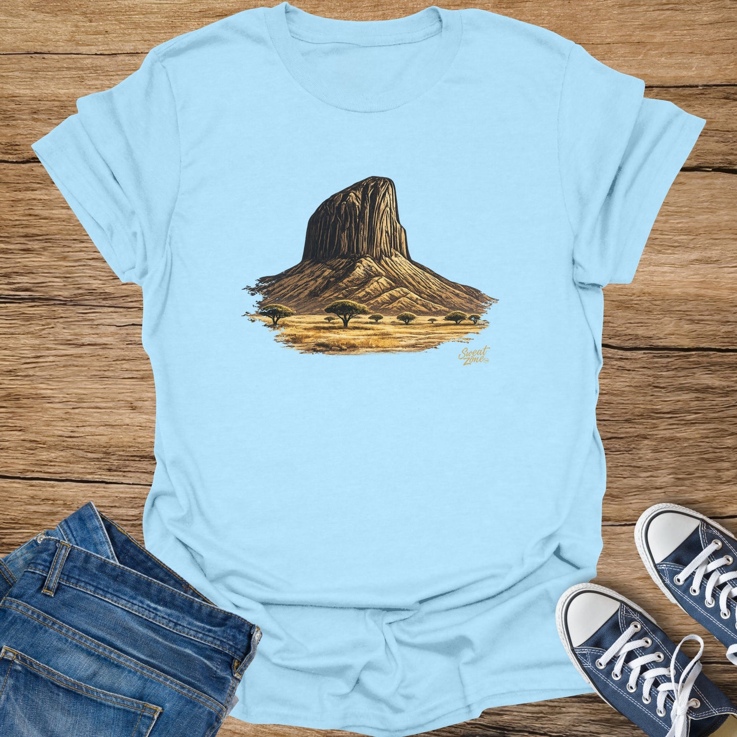 Light blue t-shirt with a brown, illustrated rocky mesa with trees on a plain.
