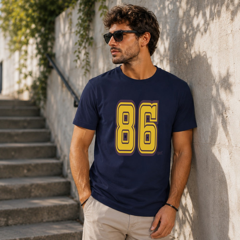 Man in navy t-shirt with yellow "86" graphic, leaning against wall.