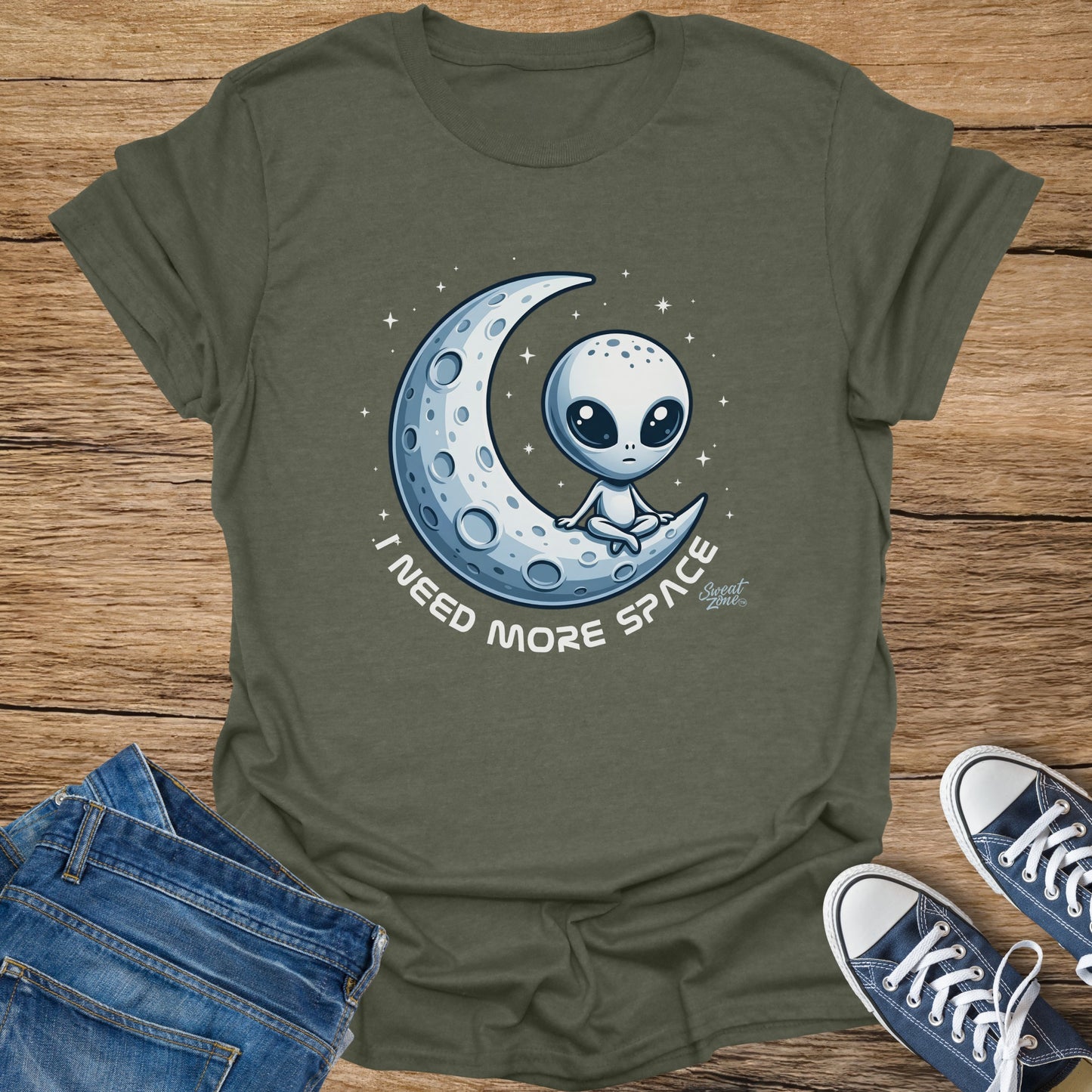 Olive t-shirt with a cartoon alien sitting on a crescent moon, text says "I NEED MORE SPACE."