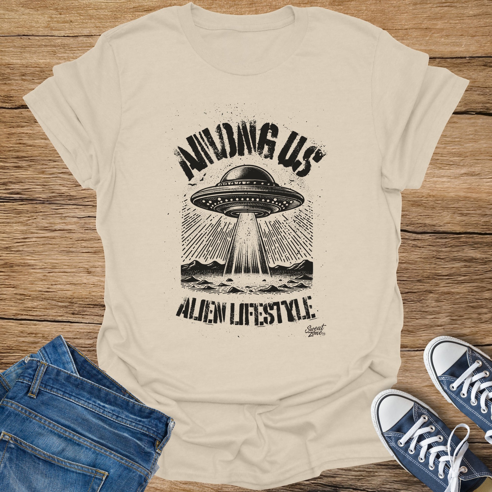 Light tan t-shirt with black graphic featuring a UFO, mountains, and text "AMONG US ALIEN LIFESTYLE".