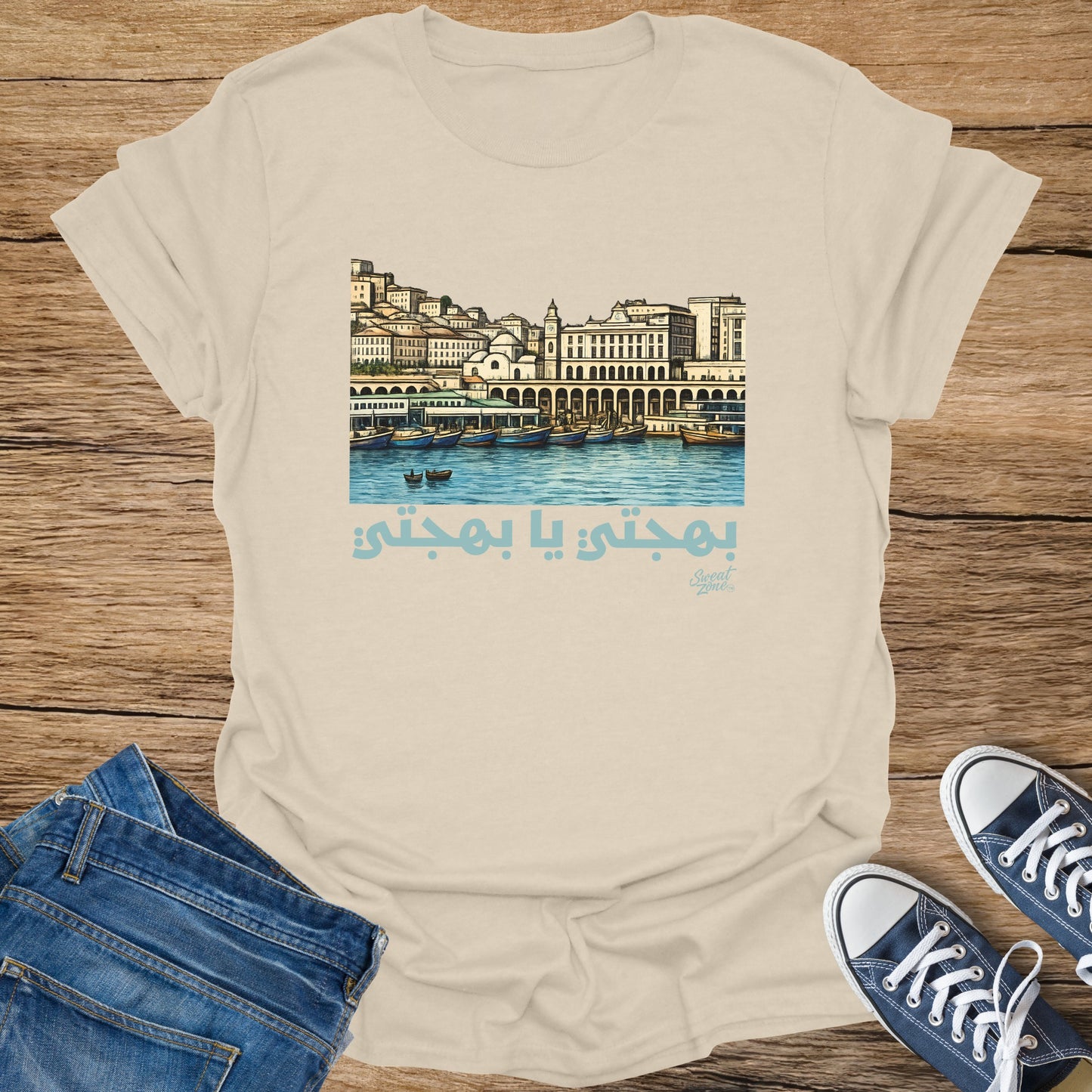 Beige t-shirt with illustration of a cityscape over water and Arabic text.