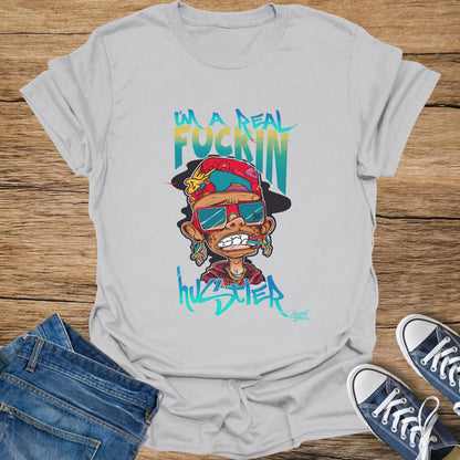 White t-shirt with a graphic of a cartoon character with "I'm a real fuckin hustler" text.