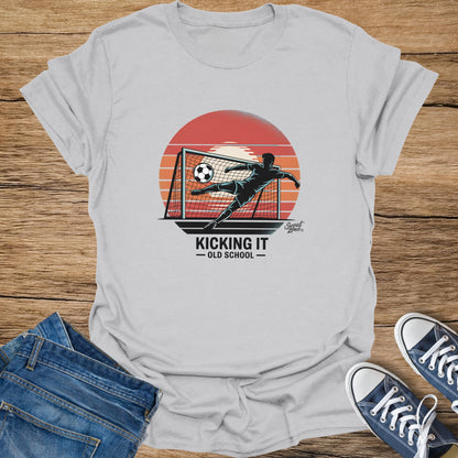 T-shirt graphic of soccer player kicking ball into a net with sunset and text "Kicking It Old School."