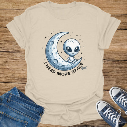 Cream t-shirt with cartoon alien sitting on crescent moon and text "I NEED MORE SPACE".